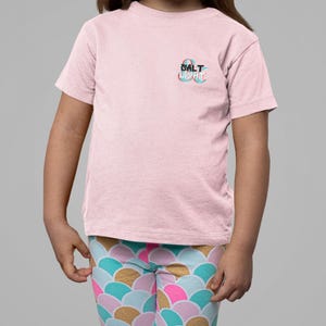 May include: A light pink t-shirt with the text "SALT & LIGHT" in a teal and blue design. The shirt is paired with leggings featuring a colorful, scale-like pattern in gold, pink, teal, and blue.