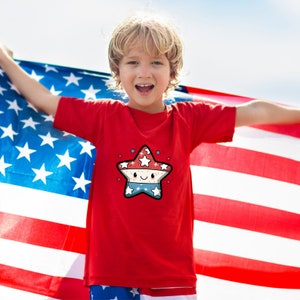May include: A red t-shirt with a cartoon star design. The star is red, white, and blue and has a smiling face. The child wearing the shirt is holding an American flag.