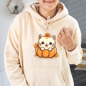 May include: A cream-colored hoodie with a cute cartoon cat wearing a pumpkin hat. The cat is sitting inside a pumpkin with orange and green leaves.