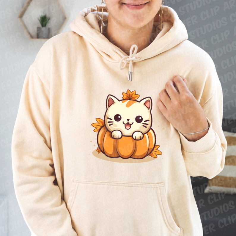 May include: A cream-colored hoodie with a cute cartoon cat wearing a pumpkin hat. The cat is sitting inside a pumpkin with orange and green leaves.