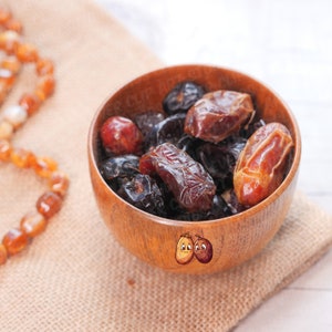 May include: A wooden bowl filled with dates. The dates are a mix of light brown and dark brown colors. The bowl is sitting on a brown burlap surface.