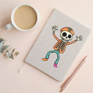 Puede incluir: Una libreta beige con un esqueleto de dibujos animados colorido que lleva un sombrero y pantalones vaqueros azules. La libreta tiene una cuerda blanca atada a ella y un bol&iacute;grafo dorado est&aacute; acostado a su lado.