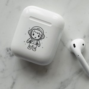 May include: White wireless charging case with a black and white illustration of an astronaut. The astronaut is wearing a helmet and a spacesuit.