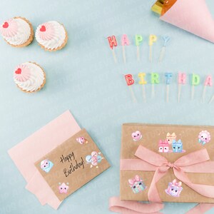 May include: A brown gift box with a pink ribbon and cute cartoon stickers. The box is surrounded by pink and white cupcakes, colourful candles that spell "Happy Birthday", and a pink and white card that says "Happy Birthday!"