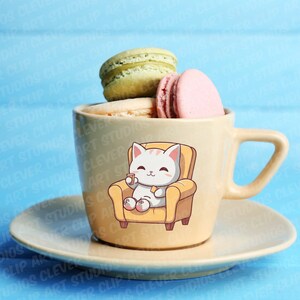 May include: A yellow ceramic mug with a cartoon cat sitting in a yellow armchair, holding a cup. The mug is filled with macarons, including green, white, and pink macarons. The mug is sitting on a matching saucer.