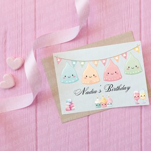 May include: A light blue card featuring a blue, yellow, pink, and green cartoon character with a happy face. The card has a banner with flags that say "Nadia's Birthday". There are also cartoon cupcakes and champagne glasses on the card.