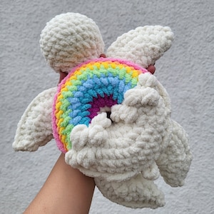 May include: A crocheted white turtle holding a rainbow-colored crocheted ring. The turtle has a fluffy texture and is a handmade toy.