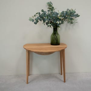 Può includere: Un tavolo a mezza luna in legno chiaro con un piano curvo e quattro gambe. Un vaso di vetro verde con steli verdi e fiori bianchi è posto sul tavolo.