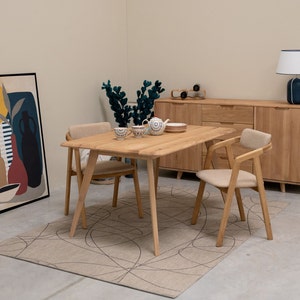 Può includere: Un tavolo da pranzo in legno chiaro con quattro sedie, un credenza e una lampada. Il tavolo ha un piano e gambe in legno chiaro, e le sedie hanno telai in legno chiaro e cuscini beige. La credenza è in legno chiaro e ha due ante. La lampada ha un paralume bianco e una base nera.