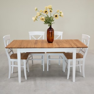 May include: A white wooden dining table with four white chairs. The table has a natural wood top and the chairs have a cross-back design. A vase with yellow flowers sits in the center of the table.