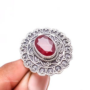 May include: A silver ring featuring a large, oval-shaped, faceted deep red gemstone at the center. The setting is circular with intricate scrollwork, beaded edges, and a dark oxidized finish. The text RhythmOfJewels is visible on the stone.