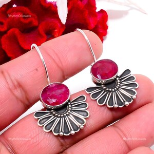 May include: A pair of silver dangle earrings featuring oval-shaped, faceted deep pink ruby gemstones. Each earring has a fan-shaped base with oxidized silver detailing and a thin silver hook for attachment. The text RhythmOfJewels is visible.