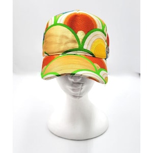 Può includere: Un cappellino da baseball colorato con un vivace motivo di forme astratte arancioni, gialle e verdi. Il cappellino è esposto su una testa di manichino bianca, mettendo in risalto il suo design. Il tessuto ha un aspetto strutturato.