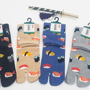 Pode incluir: Quatro pares de meias tabi em preto, bege, azul e cinza, cada um com ilustrações de sushi. As meias têm um design de dedo dividido e são exibidas com uma pequena espada e borla. Cada par está em um cabide preto com uma etiqueta.