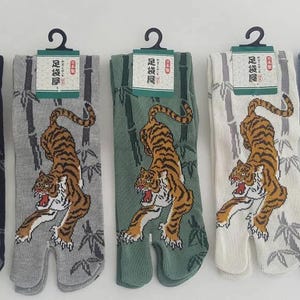 May include: Five pairs of Japanese tabi socks in various colors, each featuring a tiger design. The socks are navy, gray, green, white, and blue, with a split toe design. Each pair has a tag with Japanese text.