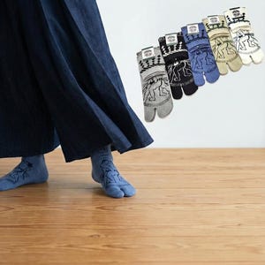 Könnte beinhalten: Eine Auswahl an Zehensocken in Grau, Schwarz, Blau und Beige, jede mit einer weißen Grafik einer Person. Die Socken werden vor einem weißen Hintergrund und einem Holzboden gezeigt.