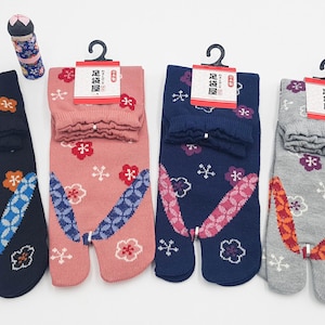 May include: Four pairs of Japanese tabi socks in navy, pink, blue, and gray, each with a split toe design. The socks feature floral patterns and decorative accents. A small, patterned decorative item is also present.
