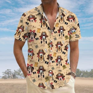 Pode incluir: Uma camisa de manga curta com botões em bege claro com uma estampa de personagens de desenhos animados usando chapéus de pirata, junto com pequenas ilustrações de âncoras, mapas do tesouro e bandeiras piratas, com os nomes Mickey, Pluto e Goofy.