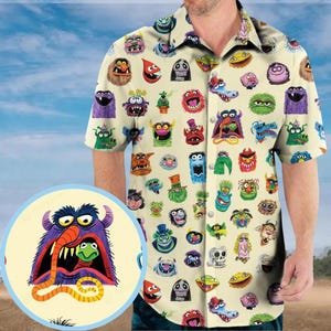 May include: Short-sleeved button-down shirt with a cream background covered in colorful cartoon monster faces. The shirt has a classic collar and button closure. The monsters have various expressions and colors.
