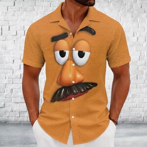 May include: A short-sleeved button-down shirt in a mustard yellow color featuring a large graphic print of a cartoon face with a prominent nose, thick black eyebrows, white eyes, and a dark mustache centered on the front of the garment.