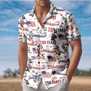Puede incluir: Una camisa blanca de manga corta con botones y un patrón repetitivo de personajes de Disney, banderas estadounidenses y el texto 250 YEARS y SEMIQUINCENTENNIAL SINCE 1776 en rojo, blanco y azul. La camisa tiene cuello y cierre frontal de botones.