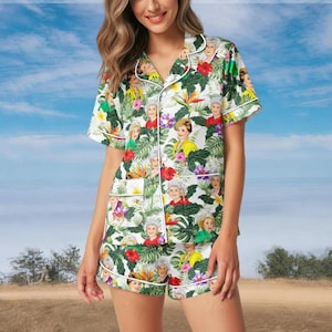 Può includere: Pigiama bianco con stampa floreale tropicale e personaggi. La maglia a maniche corte e i pantaloncini presentano un motivo di fiori colorati, foglie verdi e ritratti di donne. Il set ha bordini bianchi.