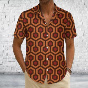 Puede incluir: Una camisa de manga corta con botones y un patrón geométrico repetitivo de hexágonos entrelazados en tonos rojo oscuro, naranja quemado y marrón. La camisa tiene un cuello clásico, una tapeta frontal con botones blancos y un dobladillo recto.