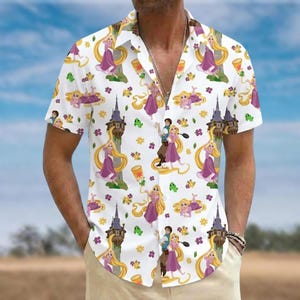 Op de afbeelding: Een wit overhemd met korte mouwen en knoopsluiting, voorzien van een all-over print met een blonde figuur met lang haar, een mannelijke figuur, hoge stenen torens, zwevende lantaarns, kleine groene kameleons en paarse en gele bloemen.