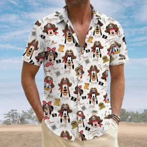 Op de afbeelding: Een wit overhemd met korte mouwen en knoopsluiting, voorzien van een all-over print van stripfiguren met piratenhoeden, waaronder Mickey Mouse, Minnie Mouse, Goofy en Pluto, afgewisseld met kleine piratenpictogrammen en cursieve tekst.