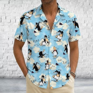 May include: A light blue short-sleeve button-down shirt featuring an all-over print of cartoon penguins in various poses, interspersed with white clouds and black musical notes. The shirt has a classic collar and a front button closure.