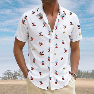 Puede incluir: Una camisa blanca de manga corta con botones y un estampado integral de Mickey Mouse con sombrero de mago azul y túnica roja. El patrón incluye pequeñas estrellas doradas y líneas azules claras en forma de remolino sobre la tela.