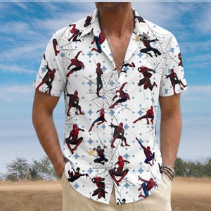 May include: White short-sleeve button-down shirt featuring a repeating pattern of Spider-Man figures, spiderwebs, and blue diamond shapes. The shirt has a classic collar and is worn open. The background shows a blue sky and a field.