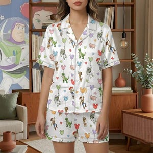 Puede incluir: Conjunto de pijama blanco con estampado de personajes de dibujos animados y globos. La parte superior de manga corta y los pantalones cortos tienen cierre de botones y bolsillos. El conjunto es de material suave y sedoso.