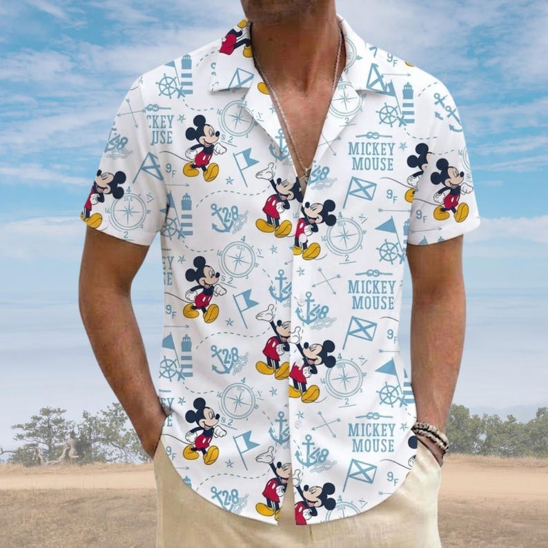 May include: White short-sleeved shirt with a repeating pattern of Mickey Mouse, anchors, compasses, and nautical flags. The shirt has the text "MICKEY MOUSE" and "CKEY JUSE" in light blue. The shirt is worn by a person.