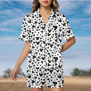 May include: White pajama set with a black Mickey Mouse pattern. The set includes a short-sleeved button-down shirt with two front pockets and matching shorts. The edges of the shirt and shorts are trimmed with white.