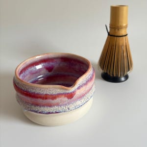 May include: A ceramic matcha bowl with a red and purple interior and a white exterior. A bamboo matcha whisk sits to the right of the bowl. The bowl has a spout for easy pouring.