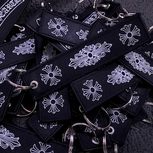 Puede incluir: Una colecci&oacute;n de llaveros rectangulares de tela negra con motivos de cruz bordados en blanco y el texto CHROME HEARTS. Cada llavero incluye una anilla de metal plateado sujeta a trav&eacute;s de un ojal met&aacute;lico reforzado en un extremo.