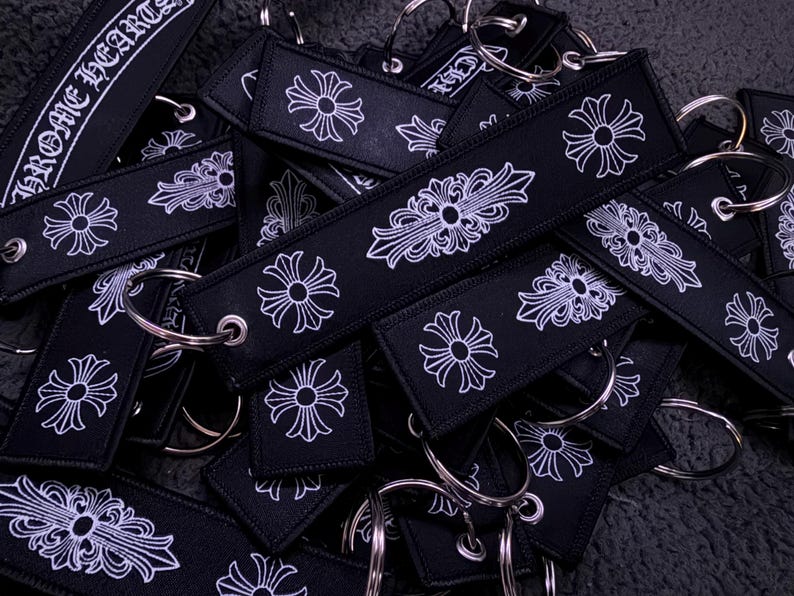 Puede incluir: Una colecci&oacute;n de llaveros rectangulares de tela negra con motivos de cruz bordados en blanco y el texto CHROME HEARTS. Cada llavero incluye una anilla de metal plateado sujeta a trav&eacute;s de un ojal met&aacute;lico reforzado en un extremo.