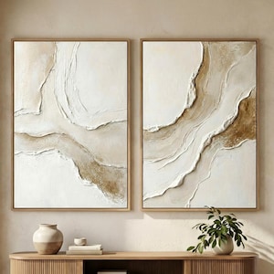 May include: Two framed abstract artworks with textured white and beige paint. The pieces feature a flowing, river-like design in a neutral colour palette. The frames are light brown. The art is displayed above a wooden cabinet.