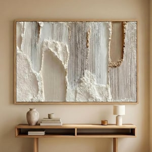 May include: Large abstract wall art in a natural wood frame. The artwork features textured white, gray, and beige elements, creating a layered, three-dimensional effect. The piece hangs above a wooden console table.