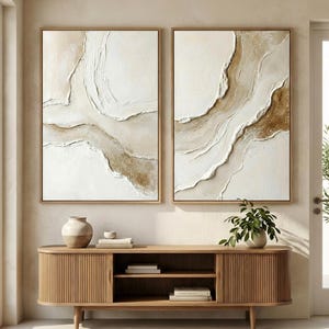 Könnte beinhalten: Zwei gerahmte abstrakte Kunstwerke mit strukturierter weißer und beiger Farbe. Die Kunstwerke zeigen fließende, geschichtete Designs in neutralen Tönen, eingefasst in hellbraunen Holzrahmen. Sie werden über einem Holzschrank ausgestellt.