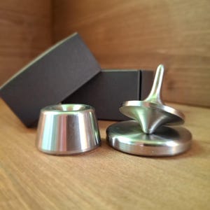 Könnte beinhalten: Ein metallischer Kreisel mit einer spitzen Stange und einer breiten, flachen Basis, begleitet von einem separaten konischen Metallständer. Die Objekte sind silberfarben mit einer glatten, polierten Oberfläche und stehen neben zwei dunkelgrauen rechteckigen Schachteln.