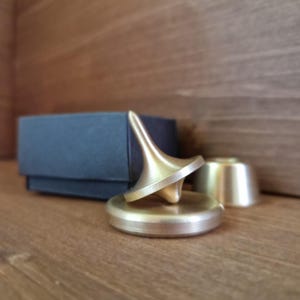 以下が含まれることがあります： 円錐形の真鍮色の独楽が円形の台座に置かれています。2つ目の独楽と小さな円筒形の部品も確認できます。木製の表面に置かれており、背景には濃い青色の箱があります。