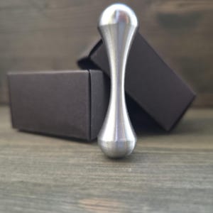 Op de afbeelding: Een glad, zilverkleurig metalen object met een slanke, zandloperachtige vorm, voorzien van een afgeronde bovenkant en een iets bredere, afgeronde basis. Het object heeft een gepolijst, reflecterend oppervlak en staat verticaal voor een donkere doos.