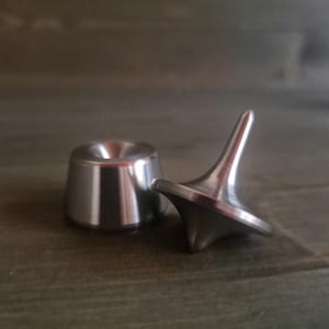 以下が含まれることがあります： 丸みを帯びたベースと尖ったトップを備えた金属製のスピニングトップと、それに合わせた金属製のベース。トップはシルバーで、滑らかで反射する表面を持っています。ベースもシルバーで、上部がくぼんでいます。