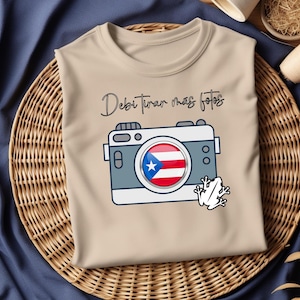 Puede incluir: Una camiseta de color beige con un gráfico de cámara y el texto "Debi tener mas fotos" en negro. La cámara tiene una bandera roja, blanca y azul dentro del objetivo y una rana blanca debajo.