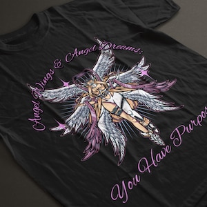 Puede incluir: Camiseta negra con un gráfico de un ángel con grandes alas blancas y moradas. El ángel lleva un atuendo blanco con detalles plateados. El texto "Angel Wings & Angel Dreams: You Have Purpose" está en fuente morada curva.