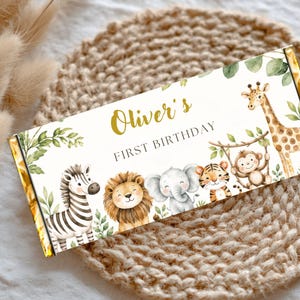 May include: A rectangular chocolate bar wrapper featuring a white background with green foliage and watercolor illustrations of a zebra, lion, elephant, tiger, monkey, and giraffe. The text reads Oliver's FIRST BIRTHDAY in gold and black.