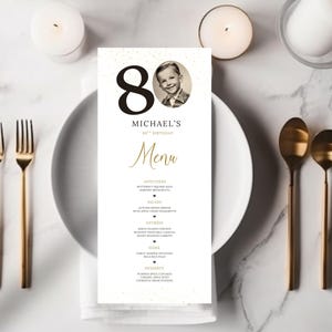 Peut inclure: Une carte de menu d'anniversaire rectangulaire affichant le nombre 80 en grande police noire, un portrait circulaire en noir et blanc d'un enfant et le texte Michael's 80th Birthday Menu. La carte liste divers plats en texte doré et noir.