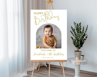 Może przedstawiać: Biały znak urodzinowy z złotym łukiem i napisem "Happy Birthday" w złotym stylu pisma. Znak zawiera również zdjęcie małego chłopca w żółtej koszuli i napis "Witamy na 1. urodzinach Michała, 14 marca 2022".